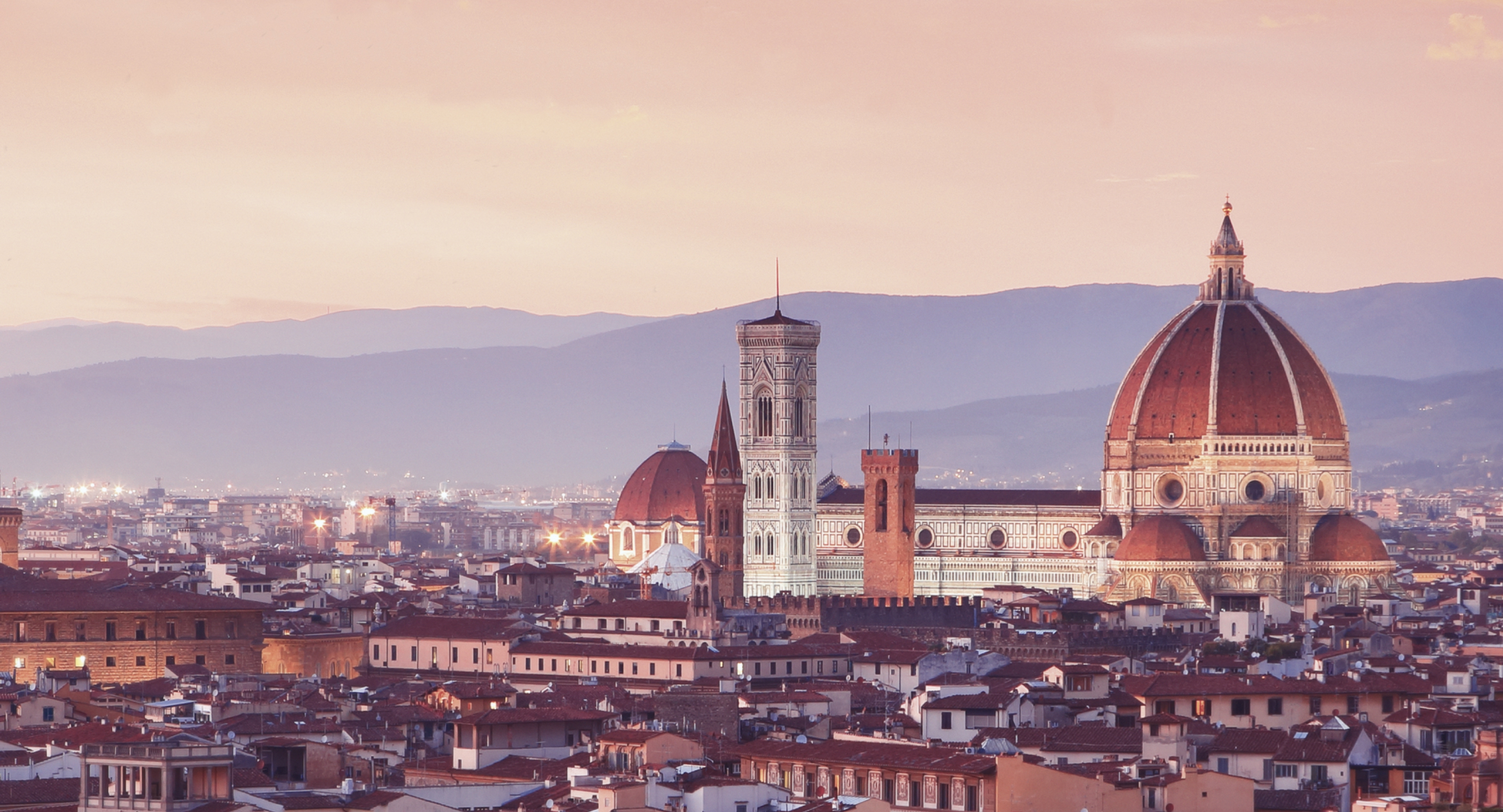 Panoramic view of Florence, Italy, at sunset featuring the Cathedral of Santa Maria del Fiore with its red-tiled dome and Giotto's Bell Tower rising above the city rooftops.