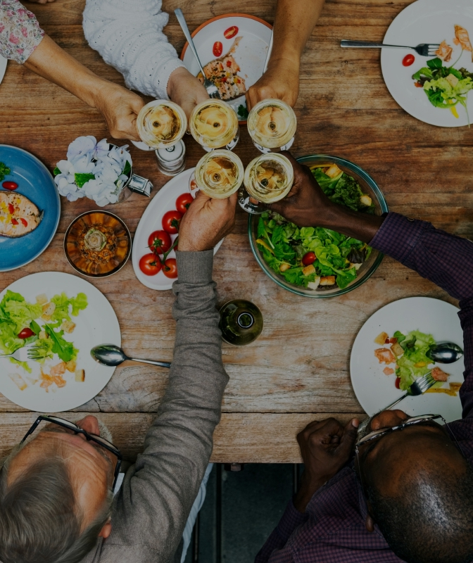Group of friends sharing a meal and toasting with drinks around a rustic wooden dining table filled with colorful salads and dishes.