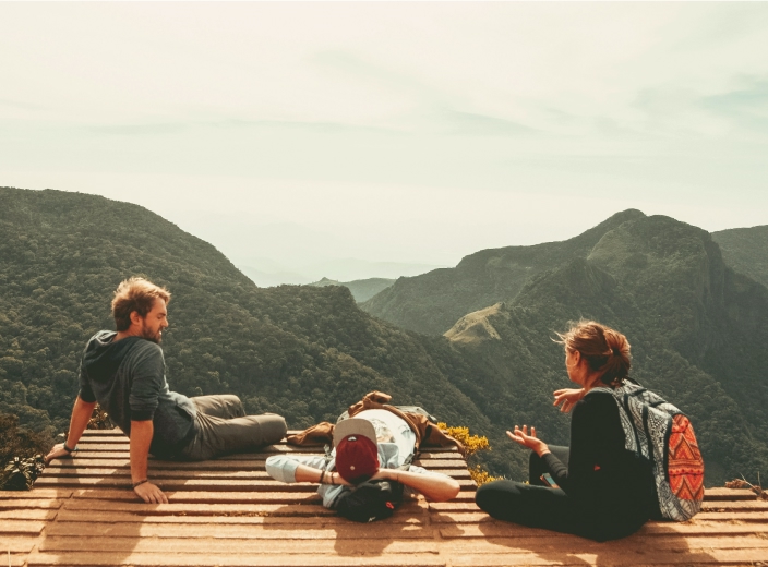 Group of friends sitting on a wooden platform overlooking a lush mountain valley under a clear sky.