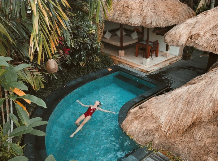 Traveler relaxing on their back in a tropical infinity pool surrounded by lush greenery and thatched-roof villas.