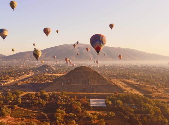 Hot air balloons floating over the ancient pyramids of Teotihuacan at sunrise, with mountains in the background.