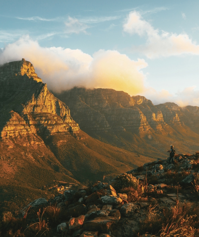 Hiker standing on a rocky trail at sunrise overlooking rugged mountain peaks and rolling clouds in Table Mountain National Park, South Africa.
